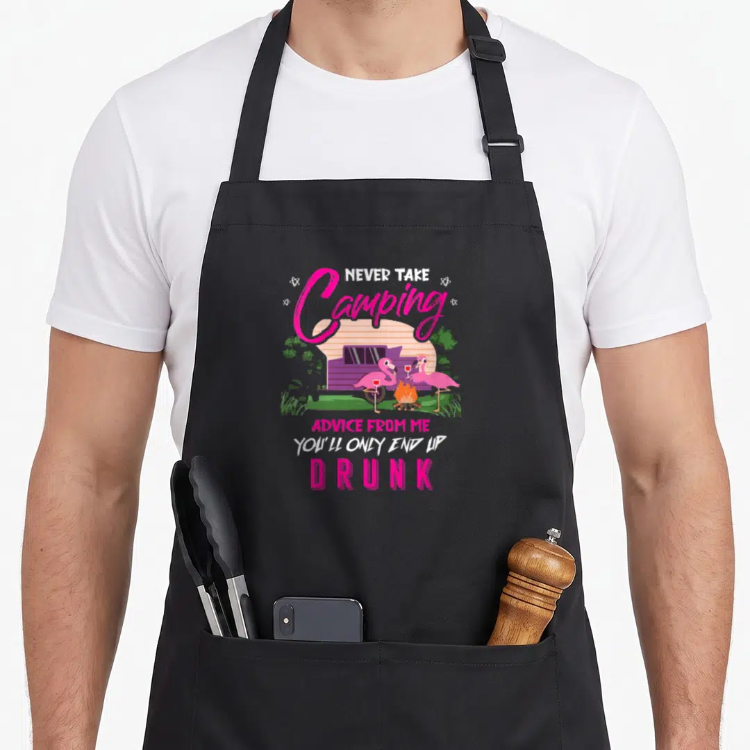 Youll camping apron - Two pink flamingos holding wine glasses by a campfire in front of a purple camper van with text — shown on a black cotton canvas apron against a white background
