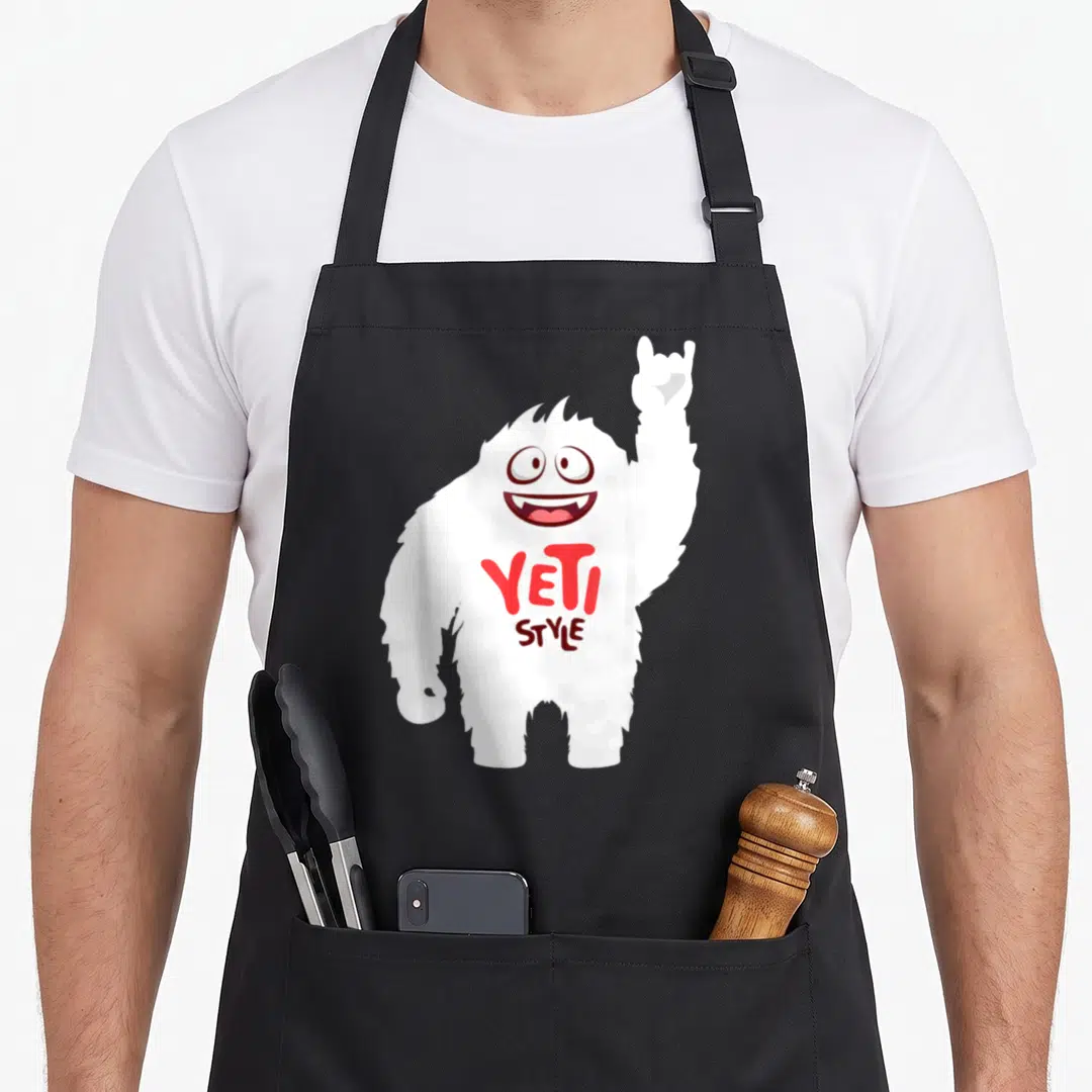 Funny yeti apron - White cartoon yeti with raised rock-on hand and YETI STYLE text in red and black — shown on a black cotton canvas apron against a white background