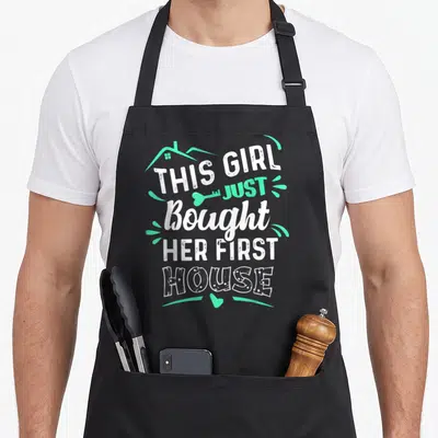 Bought owner novelty apron - White and mint green text reading THIS GIRL JUST Bought HER FIRST HOUSE with house outline — shown on a black cotton canvas apron against a white background