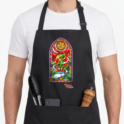 Wind stained apron - Stained glass window art of Link holding a sword with Triforce symbol at bottom — shown on a black cotton canvas apron against a white background