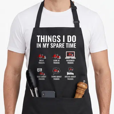Spare semi truck apron - THINGS I DO IN MY SPARE TIME text with six red truck illustrations on grey background — shown on a black cotton canvas apron against a white background