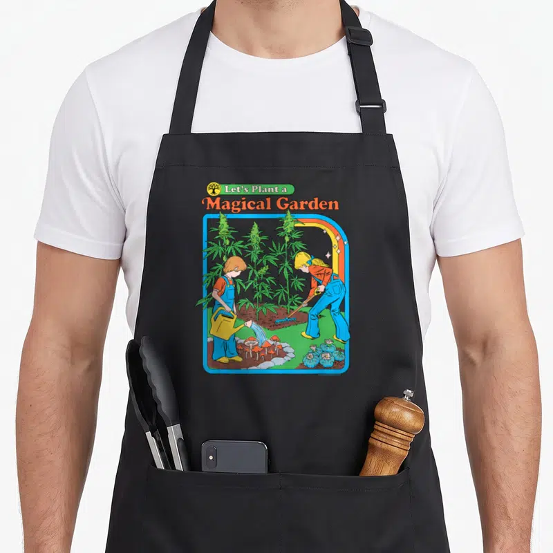 Steven mushroom apron - Two children gardening among cannabis plants and mushrooms under Let's Plant a Magical Garden — shown on a black cotton canvas apron against a white background