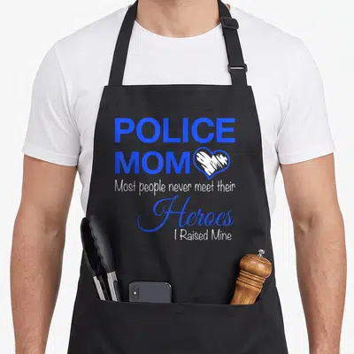 Most police apron - POLICE MOM text with white heart and blue script reading Heroes I Raised Mine — shown on a black cotton canvas apron against a white background