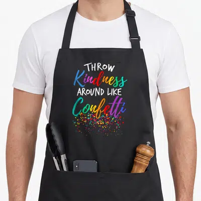 Throw teacher apron - Stacked text reading Throw Kindness Around Like Confetti with multi-colored confetti dots below — shown on a black cotton canvas apron against a white background