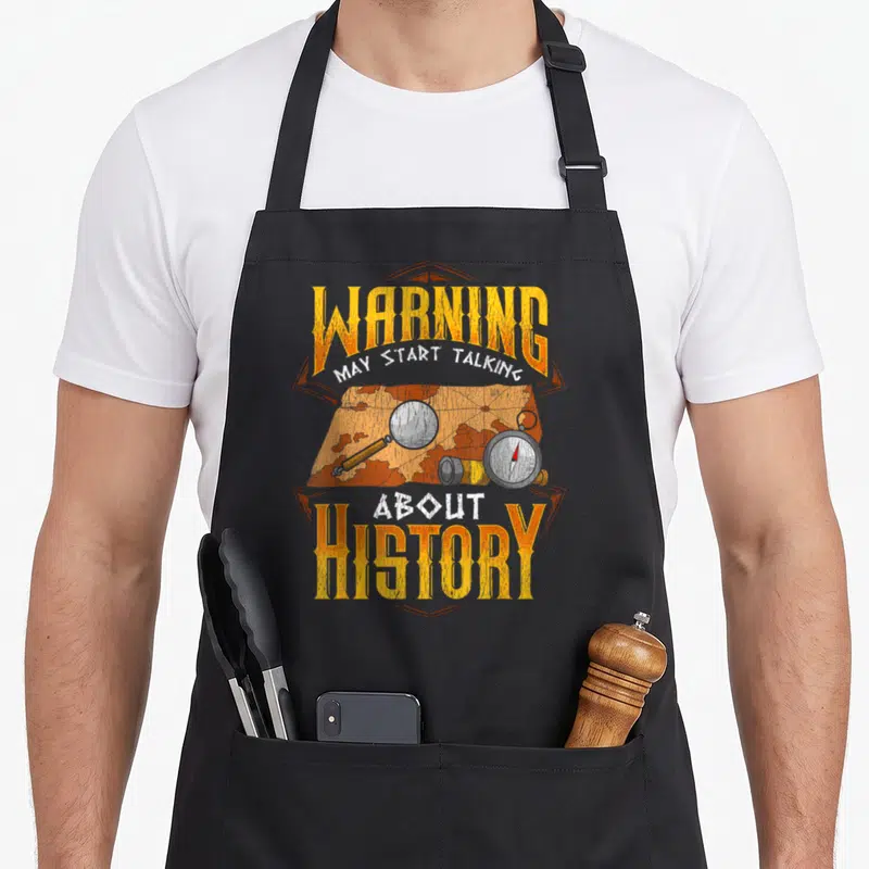 Warning historian jokes apron - Distressed map with compass, binoculars, and magnifying glass under yellow warning text — shown on a black cotton canvas apron against a white background