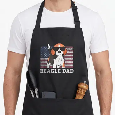 4th beagle apron - Cartoon beagle in sunglasses standing before a distressed US flag with BEAGLE DAD text below — shown on a black cotton canvas apron against a white background