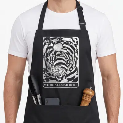 Cat mad alice wonderland apron - Cheshire Cat head in grey and white vortex with WE'RE ALL MAD HERE text — shown on a black cotton canvas apron against a white background