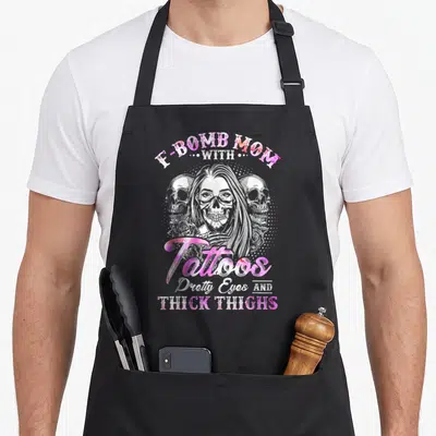 Thick tattoo apron - Skeleton woman with E-Bomb Mom text, pink lettering, two skulls and roses — shown on a black cotton canvas apron against a white background
