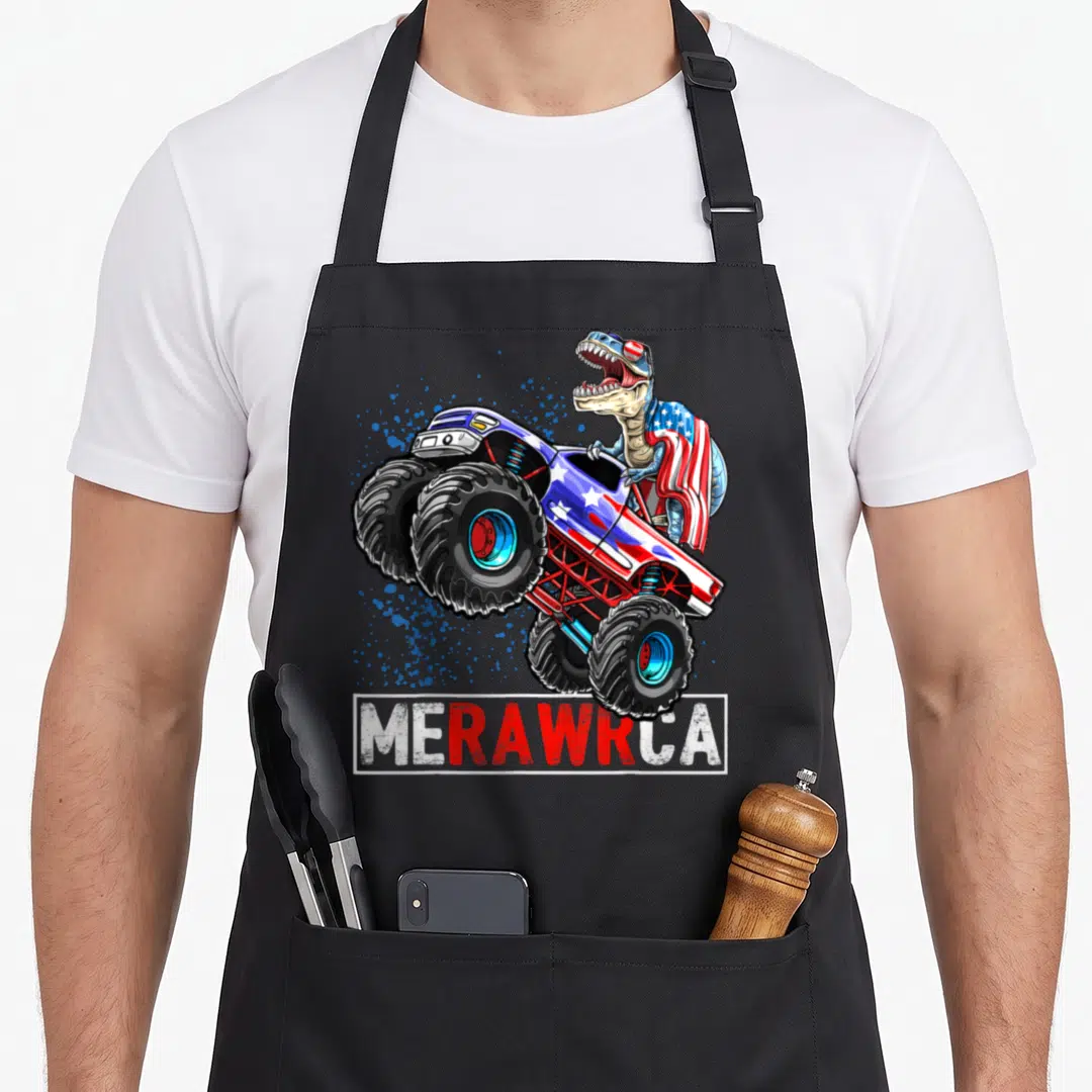 Truck 4th of july apron - T-Rex driving monster truck with Merawrca text and flag cape in blue red white beige — shown on a black cotton canvas apron against a white background