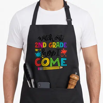 Watch 2nd apron - Stacked text reading watch out 2ND GRADE here I COME with colorful splatters and horizontal pencil — shown on a black cotton canvas apron against a white background