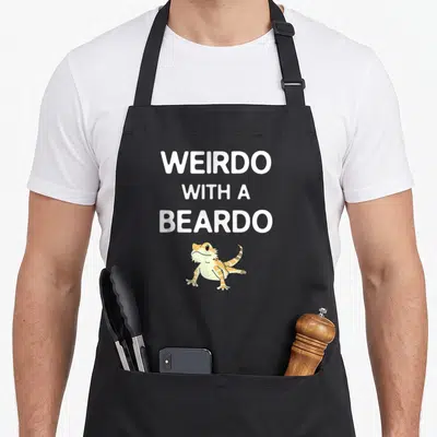 Beardo bearded dragon apron - Cartoon bearded dragon in orange and yellow below stacked white text reading WEIRDO — shown on a black cotton canvas apron against a white background
