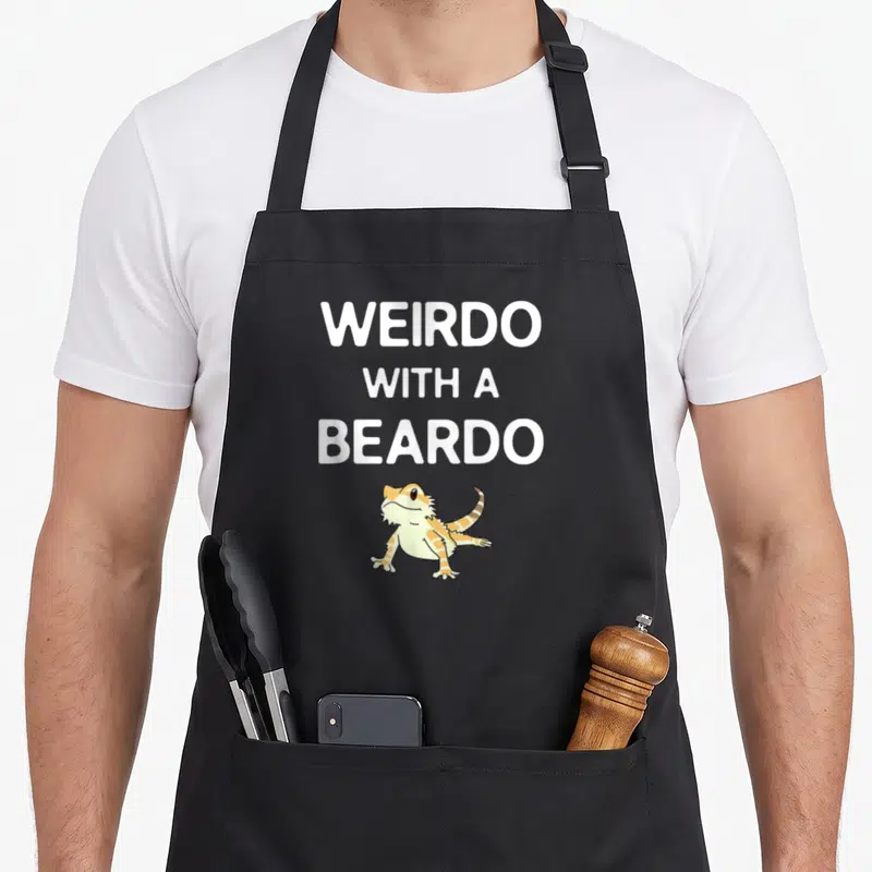 Beardo bearded dragon apron - Cartoon bearded dragon in orange and yellow below stacked white text reading WEIRDO — shown on a black cotton canvas apron against a white background
