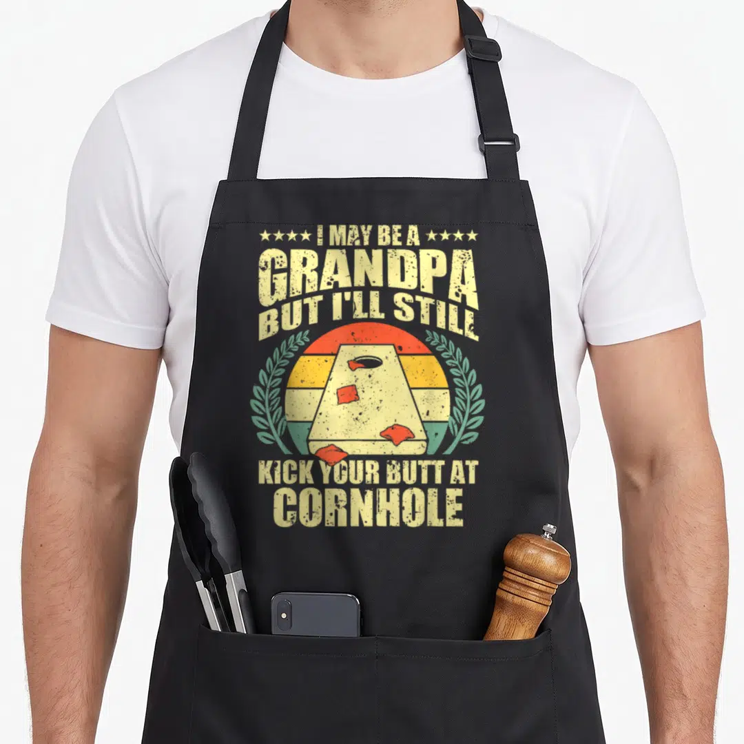 Cornhole grandpa apron - Retro sun and cornhole board with red bean bags and laurel wreaths surrounding bold text — shown on a black cotton canvas apron against a white background