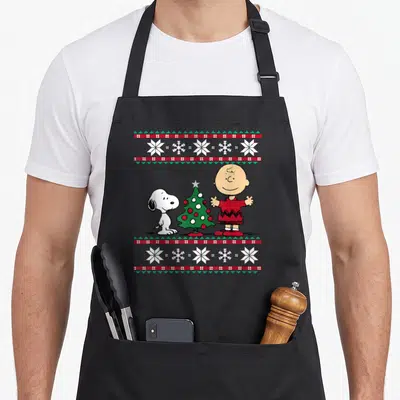 Charlie christmas tree apron - Cartoon Charlie Brown and Snoopy flanking a decorated Christmas tree with geometric — shown on a black cotton canvas apron against a white background