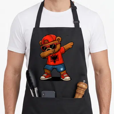 Albania bear apron - Cartoon bear dabbing in red cap and sunglasses wearing red shirt with black eagle and blue shorts — shown on a black cotton canvas apron against a white background