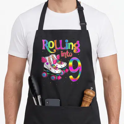 Roll turning apron - Rainbow text Rolling into 9 with pink winged roller skates and stars — shown on a black cotton canvas apron against a white background
