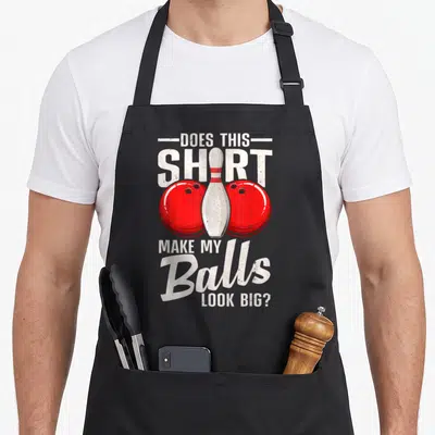 Ball bowling green apron - Distressed text DOES THIS SHIRT MAKE MY BALLS LOOK BIG with red balls and white pin — shown on a black cotton canvas apron against a white background
