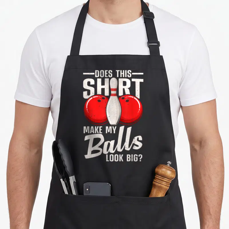 Ball bowling green apron - Distressed text DOES THIS SHIRT MAKE MY BALLS LOOK BIG with red balls and white pin — shown on a black cotton canvas apron against a white background