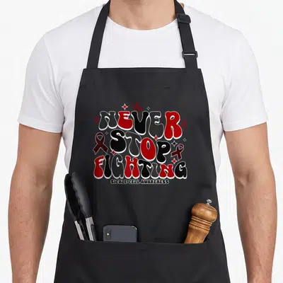 Stop sickle cell awareness apron - NEVER STOP FIGHTING text with red ribbons and butterflies in bubble font — shown on a black cotton canvas apron against a white background