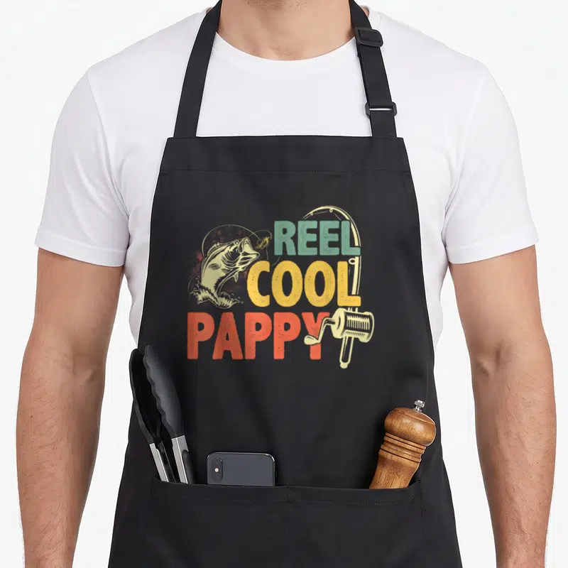 Reel pappy father day apron - Retro fishing graphic with jumping fish and REEL COOL PAPPY text in teal yellow orange red — shown on a black cotton canvas apron against a white background