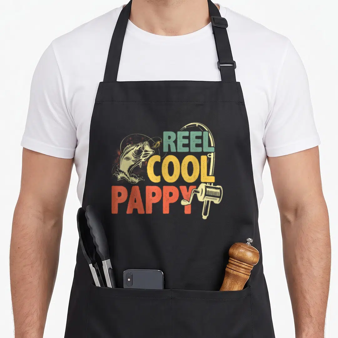 Reel pappy father day apron - Retro fishing graphic with jumping fish and REEL COOL PAPPY text in teal yellow orange red — shown on a black cotton canvas apron against a white background