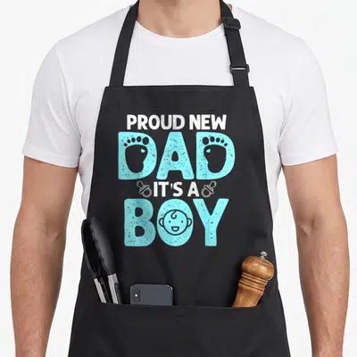 Cute proud father day apron - Stacked PROUD NEW DAD IT'S A BOY text with baby footprints inside letters and a face — shown on a black cotton canvas apron against a white background