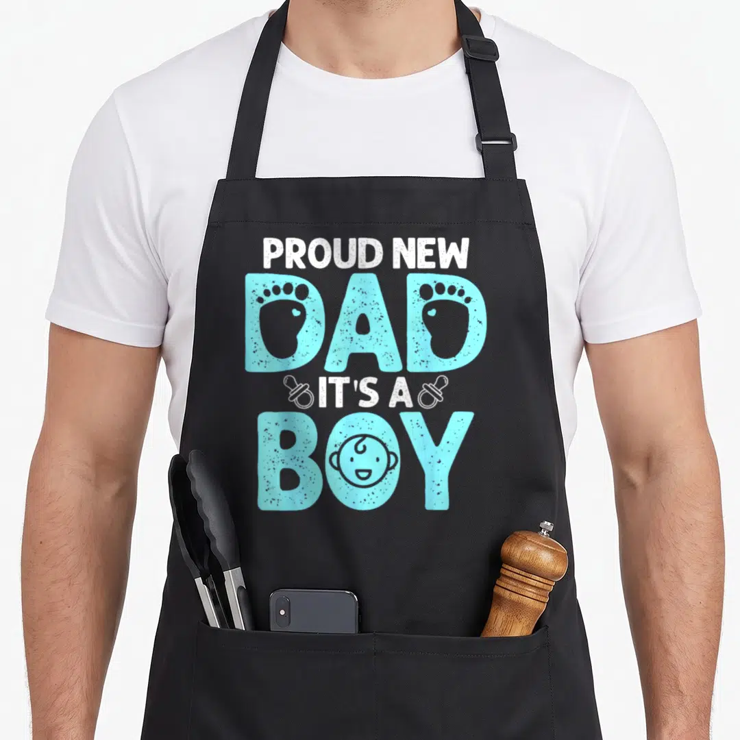 Cute proud father day apron - Stacked PROUD NEW DAD IT'S A BOY text with baby footprints inside letters and a face — shown on a black cotton canvas apron against a white background