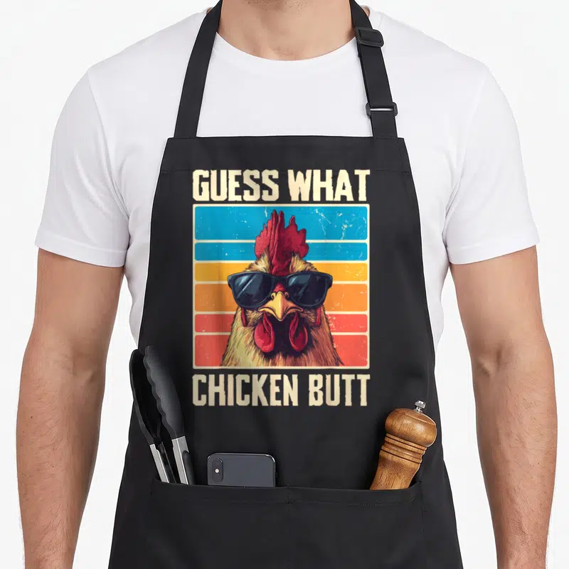 Cute guess chicken apron - Retro rooster wearing sunglasses with GUESS WHAT CHICKEN BUTT text on striped background — shown on a black cotton canvas apron against a white background