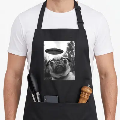 Pug selfie funny apron - Wide-eyed pug and UFO hovering above head in a black and white forest photo — shown on a black cotton canvas apron against a white background
