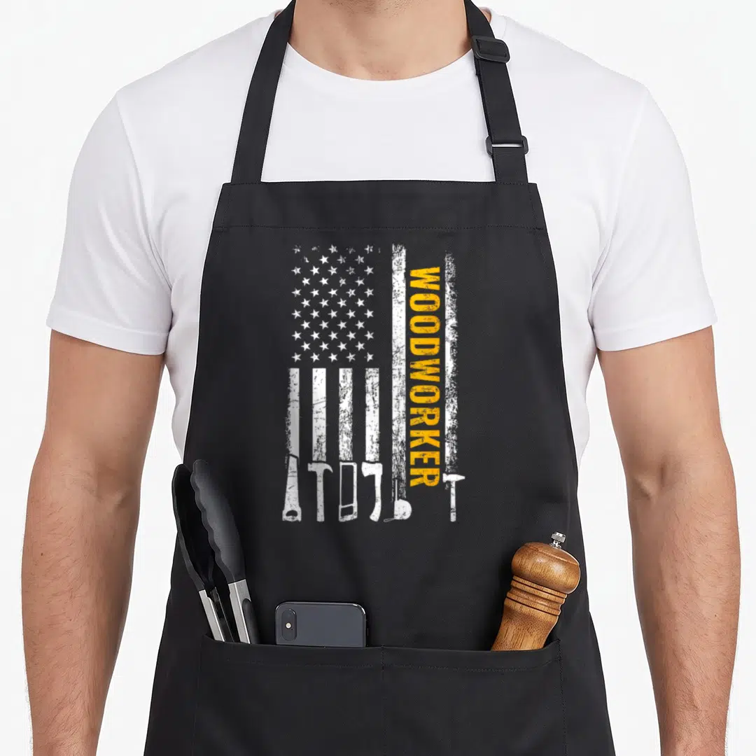 Woodworker american flag apron - Vertical orange WOODWORKER text beside distressed flag with white tool silhouettes — shown on a black cotton canvas apron against a white background