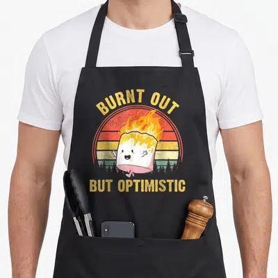 Burnt camping apron - Flaming marshmallow cartoon with arched BURNT OUT text and straight BUT OPTIMISTIC text — shown on a black cotton canvas apron against a white background