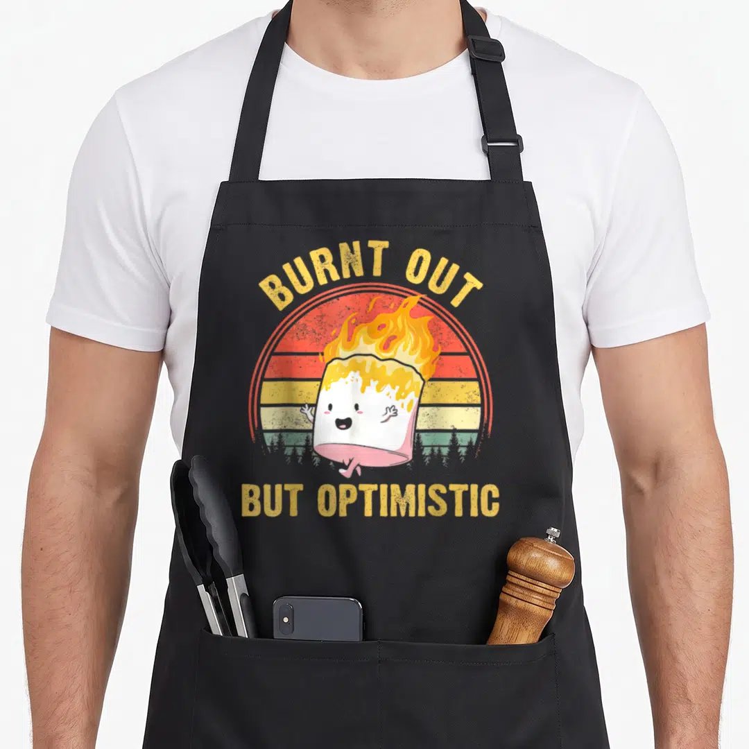 Burnt camping apron - Flaming marshmallow cartoon with arched BURNT OUT text and straight BUT OPTIMISTIC text — shown on a black cotton canvas apron against a white background