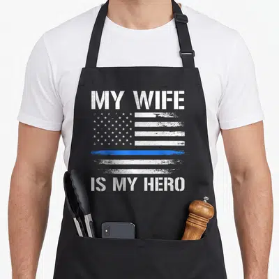 Police husband wife aprons - Distressed white and blue flag with My Wife Is My Hero text — shown on a black cotton canvas apron against a white background