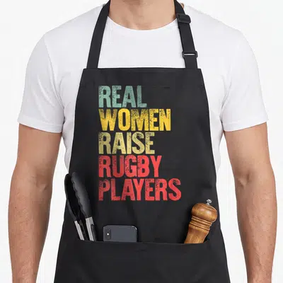 Proud rugby life apron - Stacked distressed text Real Women Raise Rugby Players in teal, yellow, olive, green, and red — shown on a black cotton canvas apron against a white background