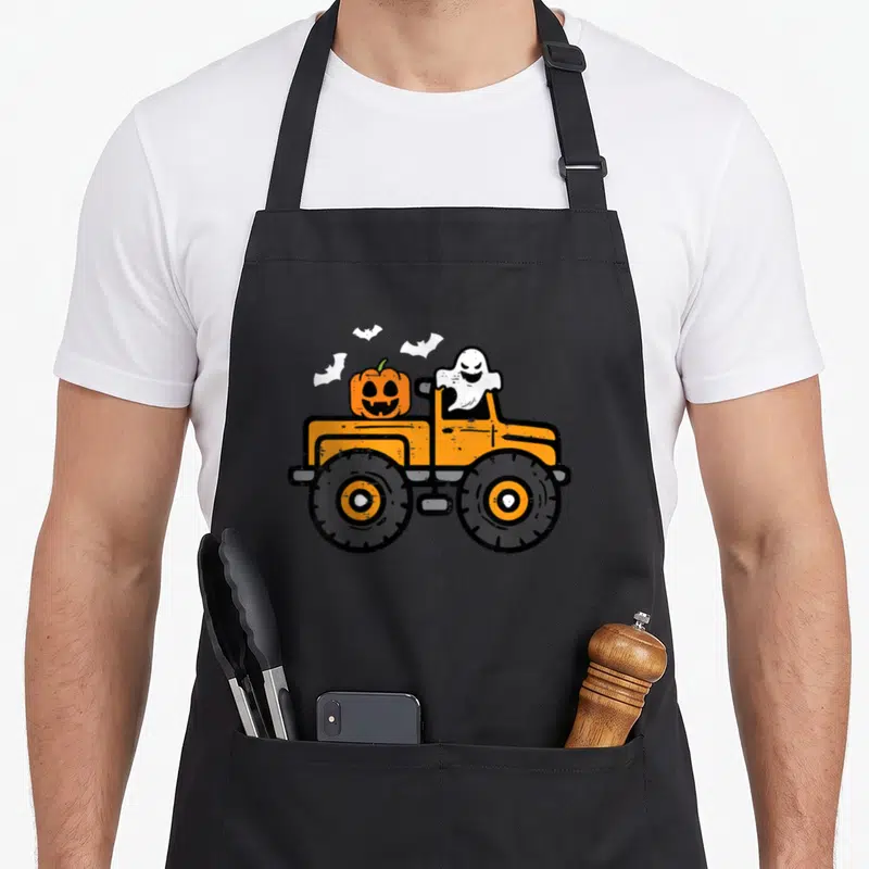 Monster pumpkin apron - Orange monster truck with white ghost driver and pumpkin cargo flanked by three bats — shown on a black cotton canvas apron against a white background
