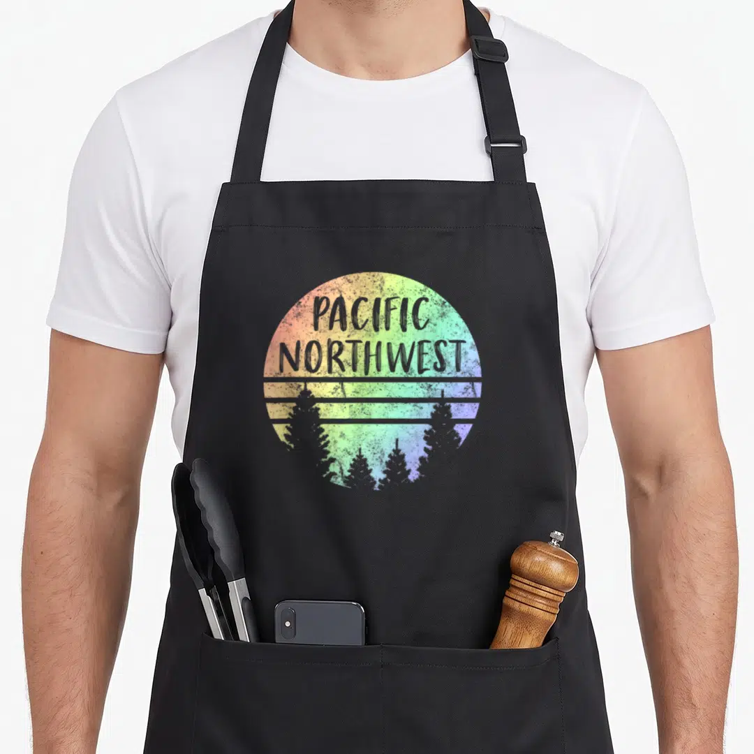 Pacific rainbow apron - Retro circle with PACIFIC NORTHWEST text, three lines, and pine trees in rainbow colors — shown on a black cotton canvas apron against a white background