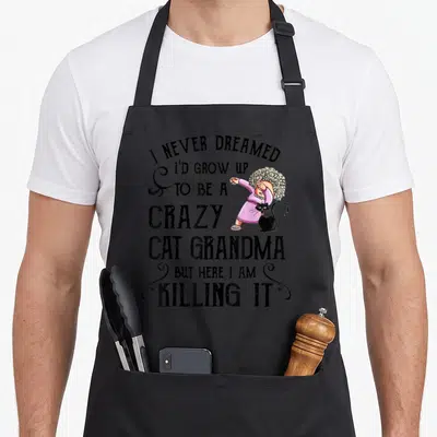 Dreamed black cat aprons - Cartoon grandma in pink dress dabbing next to a black cat with stacked humorous text — shown on a black cotton canvas apron against a white background