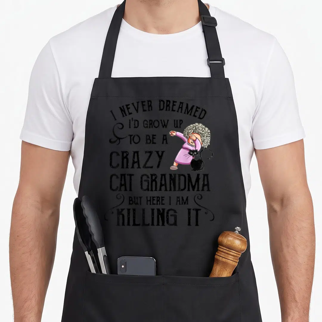 Dreamed black cat aprons - Cartoon grandma in pink dress dabbing next to a black cat with stacked humorous text — shown on a black cotton canvas apron against a white background