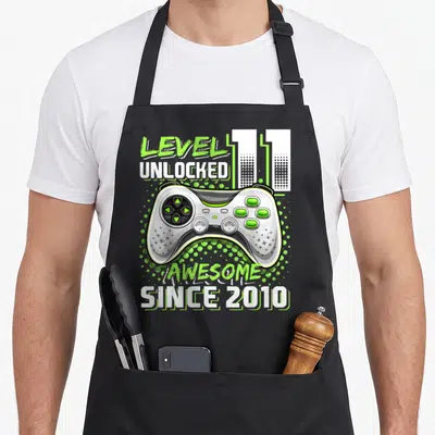 Unlocked 11th video game apron - Game controller with lime green dots and text reading LEVEL UNLOCKED 11 AWESOME SINCE — shown on a black cotton canvas apron against a white background
