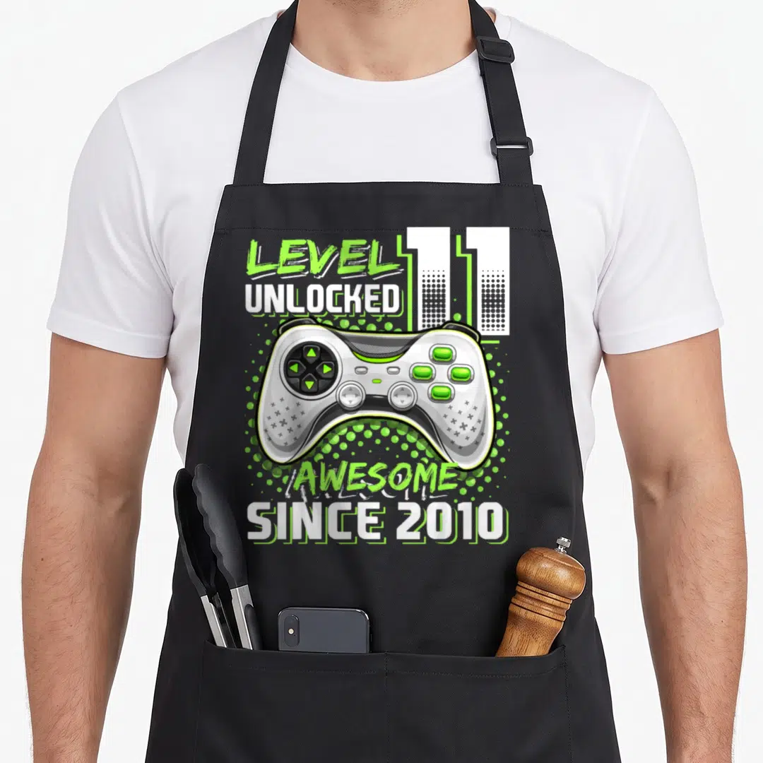 Unlocked 11th video game apron - Game controller with lime green dots and text reading LEVEL UNLOCKED 11 AWESOME SINCE — shown on a black cotton canvas apron against a white background