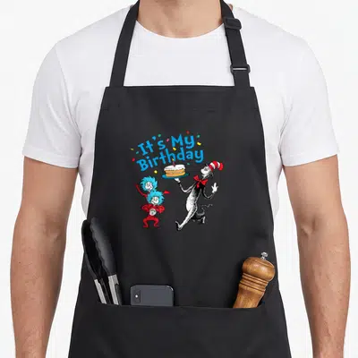 Funny general birthday apron for grilling - Cat in the Hat holds cake flanked by Thing 1 and Thing 2 under arched blue — shown on a black cotton canvas apron against a white background