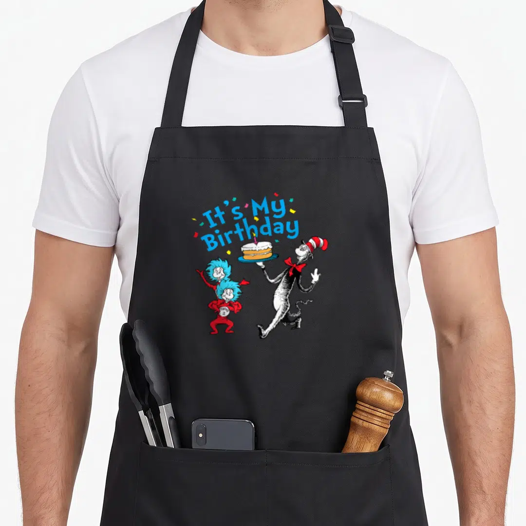 Funny general birthday apron for grilling - Cat in the Hat holds cake flanked by Thing 1 and Thing 2 under arched blue — shown on a black cotton canvas apron against a white background