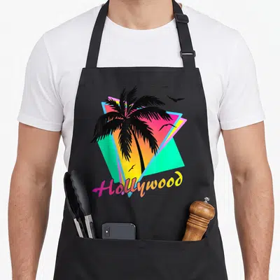 Hollywood tree apron - Black palm tree over pink, yellow, cyan triangles with three birds and Hollywood text — shown on a black cotton canvas apron against a white background