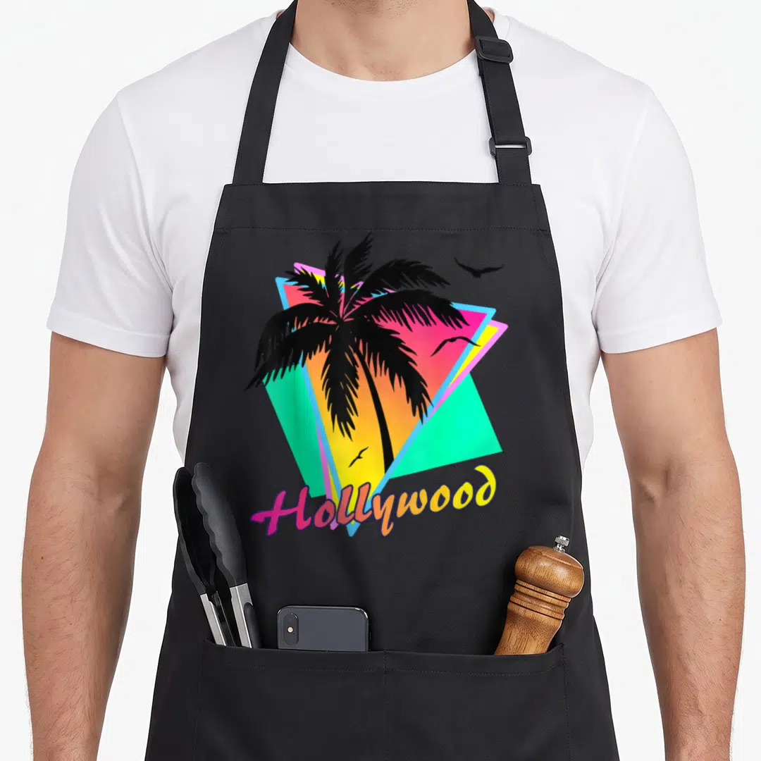Hollywood tree apron - Black palm tree over pink, yellow, cyan triangles with three birds and Hollywood text — shown on a black cotton canvas apron against a white background