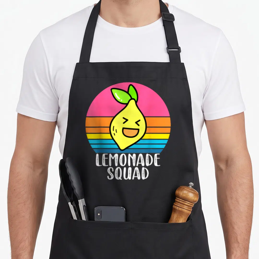 Selling lemonade apron - Cartoon lemon face over pink blue and yellow striped circle with white text — shown on a black cotton canvas apron against a white background