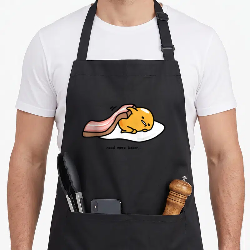 Gudetama bacon apron - Yellow egg character on white base under pink bacon strip with centered black text — shown on a black cotton canvas apron against a white background