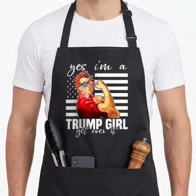 Trump aye apron - Cartoon Rosie the Riveter flexing arm with Trump Girl text and flag background — shown on a black cotton canvas apron against a white background