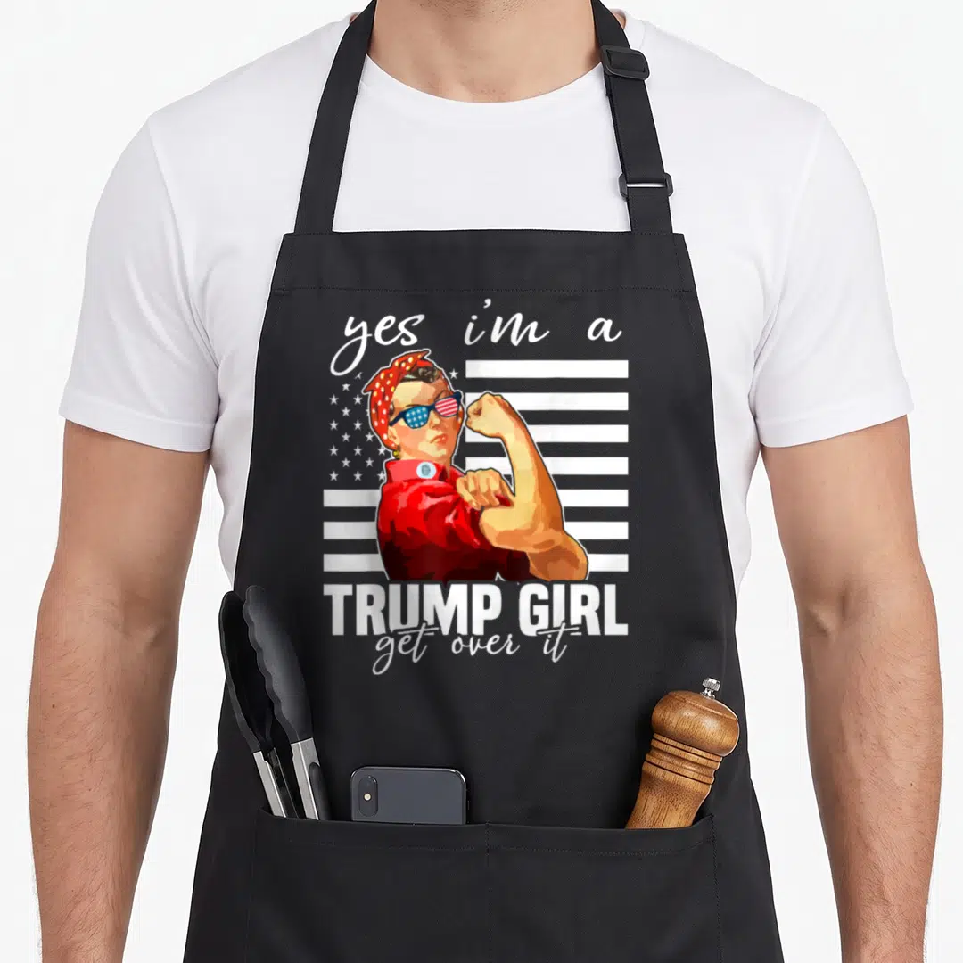 Trump aye apron - Cartoon Rosie the Riveter flexing arm with Trump Girl text and flag background — shown on a black cotton canvas apron against a white background