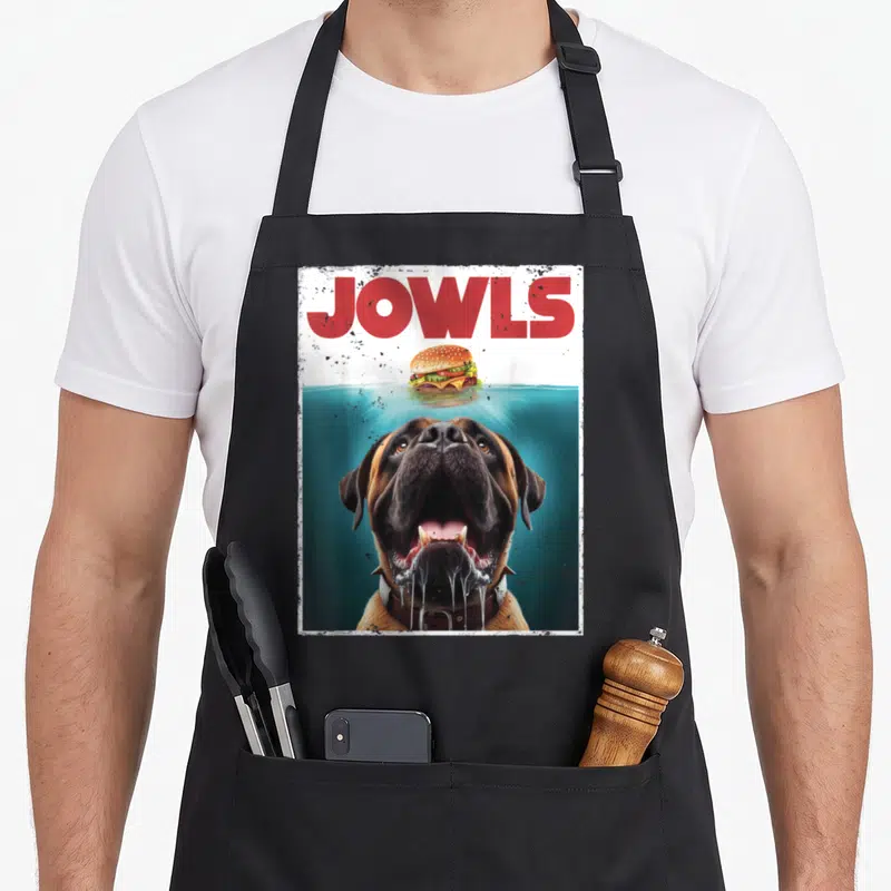 Bullmastiff dog apron - Mastiff head in teal water looking at a burger under red JOWLS text with distressed border — shown on a black cotton canvas apron against a white background