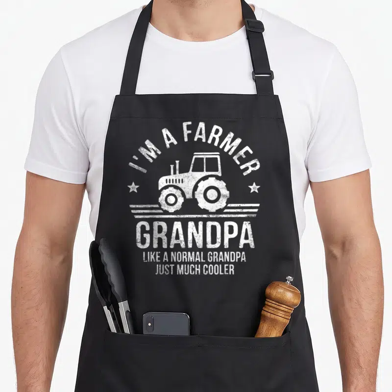 Grandpa farm apron - White distressed tractor and farmer grandpa text on black bib apron — shown on a black cotton canvas apron against a white background
